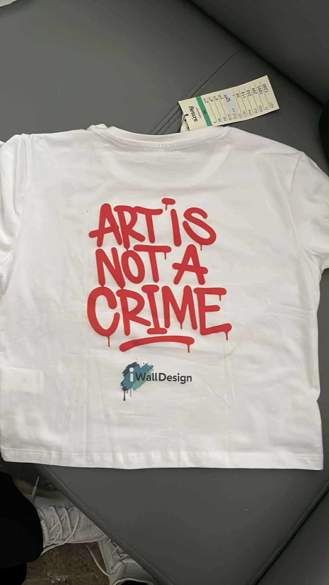 Biała koszulka z czerwonym napisem 'Art is not a crime' i logo 'iWallDesign', leżąca na szarym tle. Widoczna metka. Nietypowa czcionka i układ.