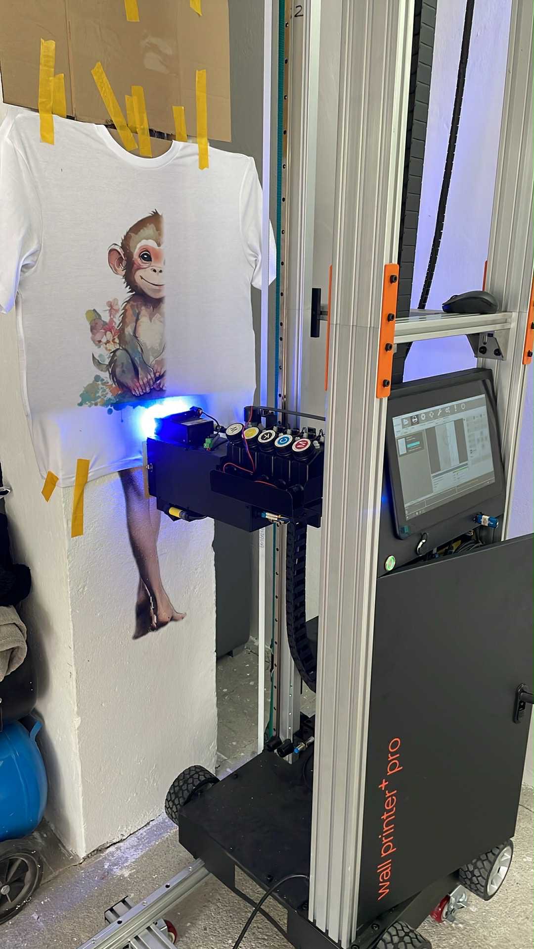 Biała koszulka z nadrukiem małpki, przymocowana do ściany taśmą. Drukarka do nadruków na odzieży podczas pracy, widoczne kartridże z kolorami i niebieskie światło.