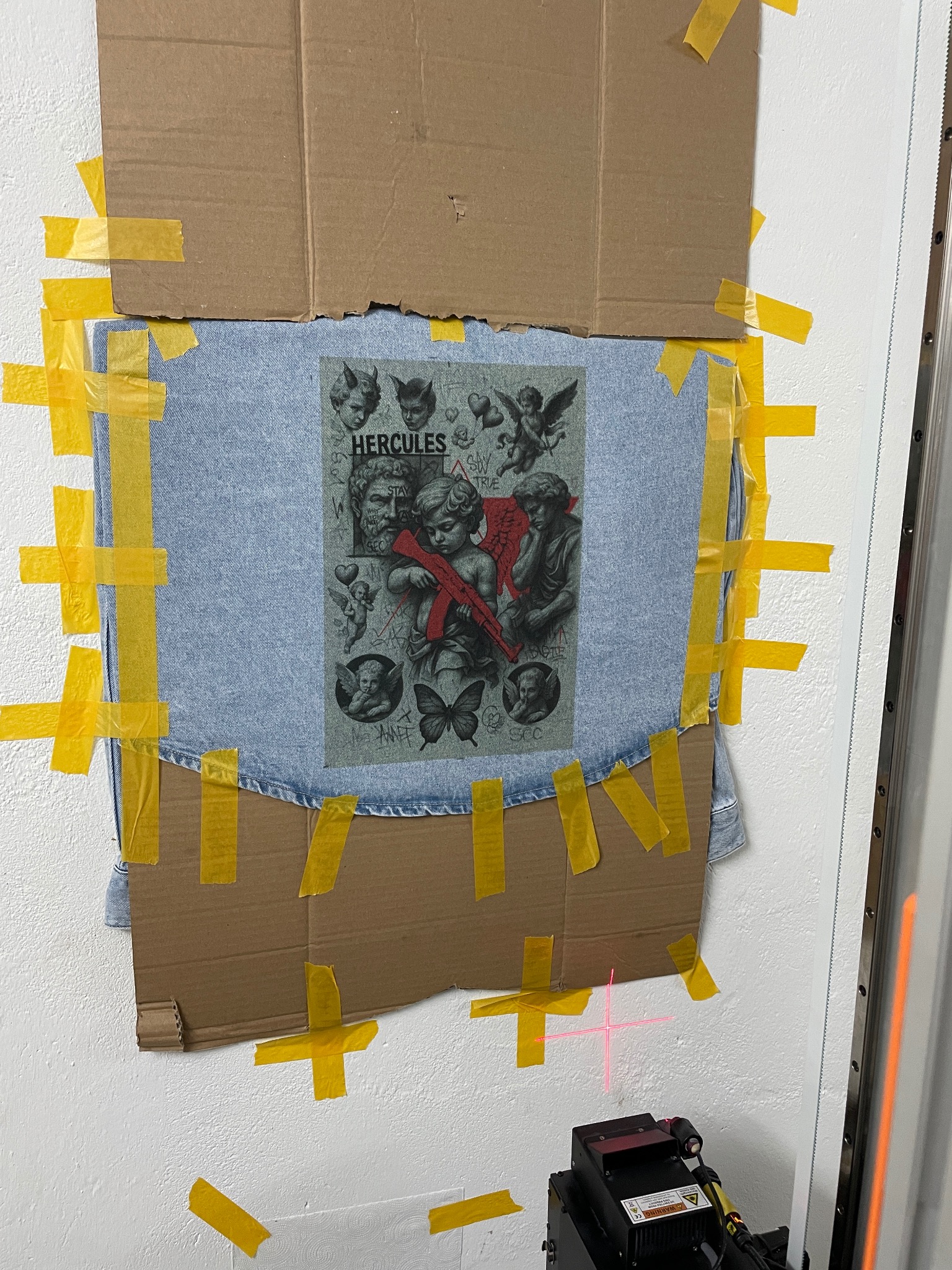 Jasna kurtka dżinsowa z nadrukiem aniołów i postaci Herculesa, przymocowana taśmą do ściany, przygotowana do dalszej obróbki przy użyciu lasera.