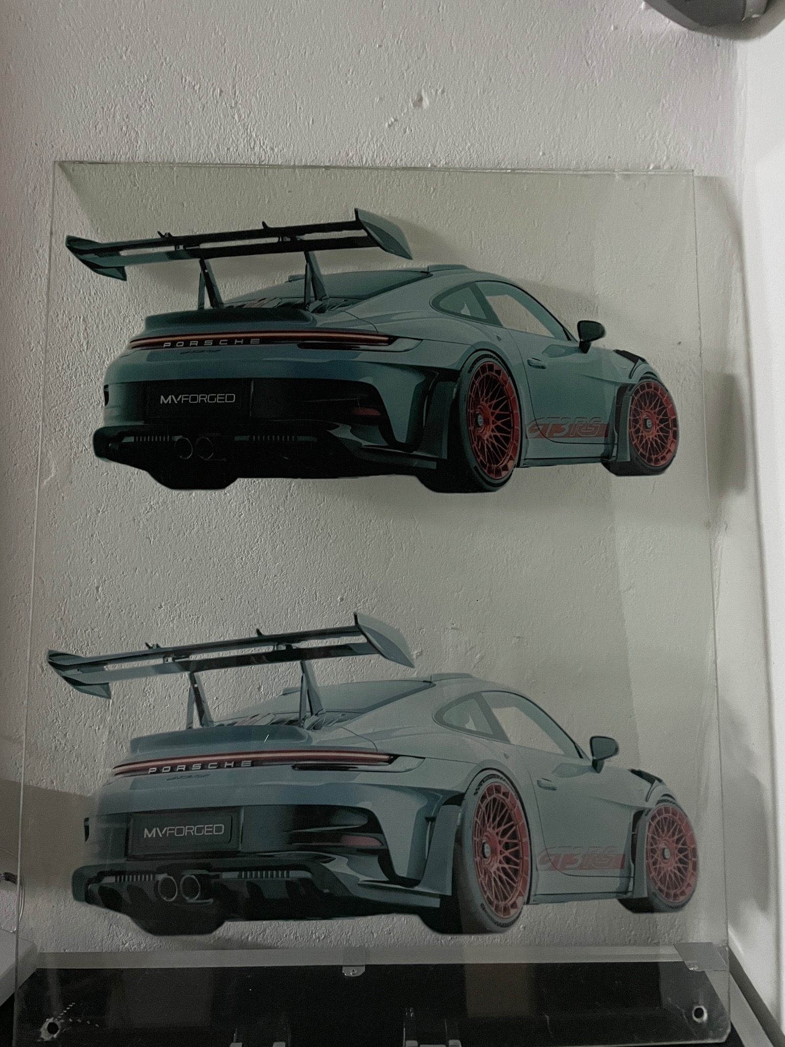 Grafika na szkle prezentuje dwa ujęcia sportowego samochodu Porsche z dużym spojlerem, z tyłu, z czerwonymi felgami, na jasnym tle ściany.