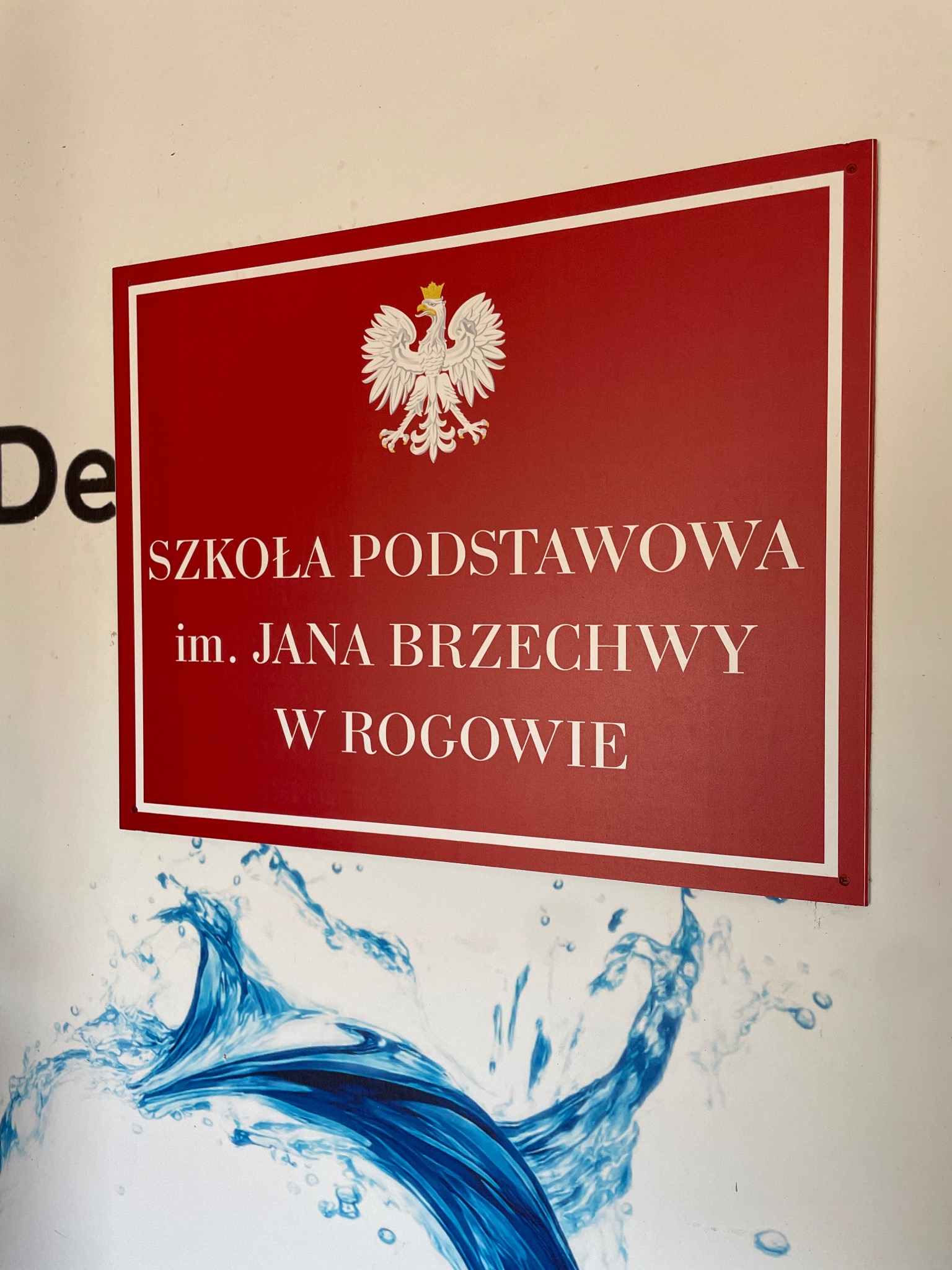 Czerwona tablica informacyjna z godłem Polski i napisem 'Szkoła Podstawowa im. Jana Brzechwy w Rogowie', umieszczona na ścianie z niebieskim motywem wodnym.