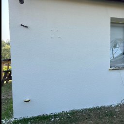 Usługi ogólnobudowlane Krystian Rakuś - Biała, świeżo otynkowana ściana zewnętrzna domu z widocznymi elementami instalacji i zabezpieczonym oknem. Tekstura tynku barankowego.