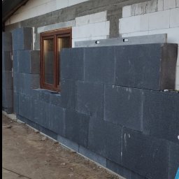 Usługi ogólnobudowlane Krystian Rakuś - Ocieplanie budynku: ciemne bloki styropianu układane na ścianie z białego bloczka, okno w drewnianej ramie, poziomica na górze. Prace w toku.