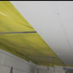 Usługi ogólnobudowlane Krystian Rakuś - Częściowy montaż sufitu z płyt karton-gips, zabezpieczony żółtą folią ochronną przy ścianie z bloczków betonowych. Widoczne łączenia płyt i przewód.