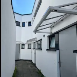 Białe budynki w wąskim przejściu, z szarymi drzwiami i daszkami, widok z dołu w kierunku nieba. Minimalistyczna architektura z wyraźnymi liniami i kątami.