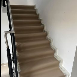 Usługi ogólnobudowlane Krystian Rakuś - Wykończone schody z beżową wykładziną, zabezpieczone taśmą malarską przy ścianie i poręczy. Widok z góry na dół, skupienie na detalu wykończenia.