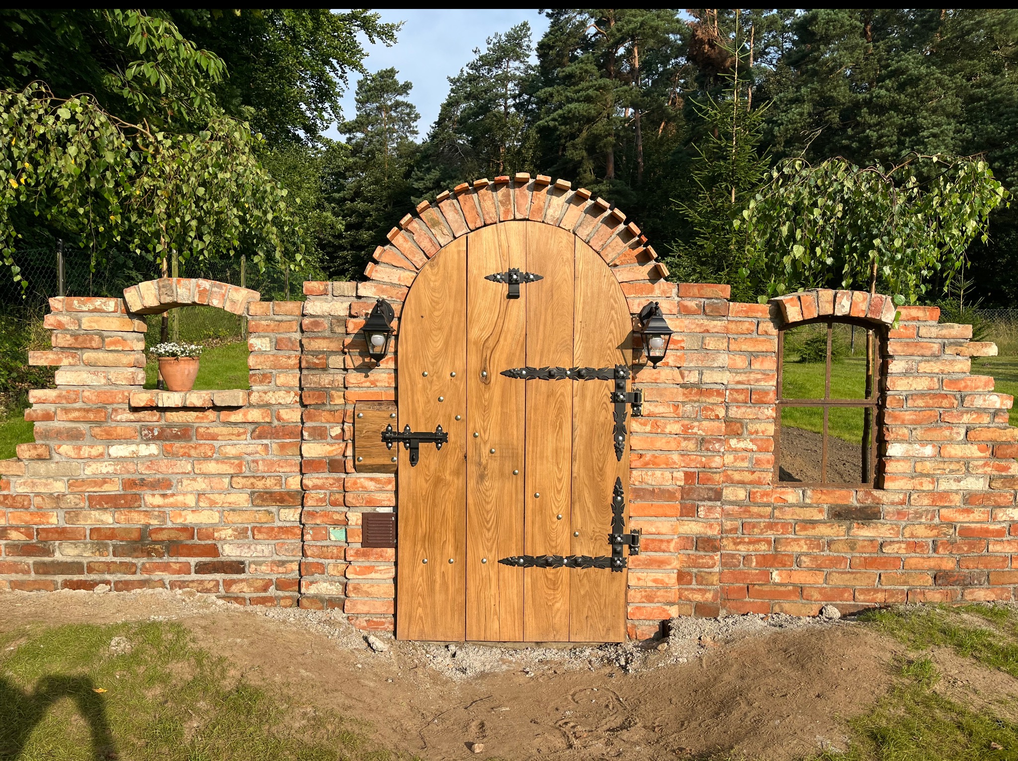 Rustykalna brama z jasnego drewna, zdobiona kutymi elementami, wkomponowana w ceglany mur z łukowymi prześwitami i lampami, otoczona zielenią.