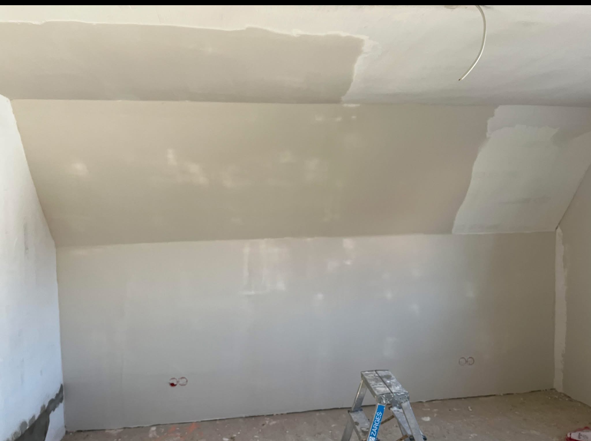 Poddasze po szpachlowaniu, widoczne gładkie ściany i częściowo wyszpachlowany sufit. Na podłodze i ścianach widoczne ślady po pracy. Aluminiowa drabina stoi w rogu.