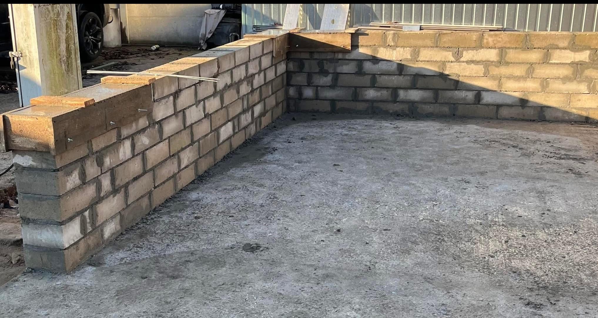 Murowana ściana z pustaków betonowych, częściowo wykończona, z elementami drewnianymi na górze, na betonowej posadzce. Widoczne ślady zaprawy.