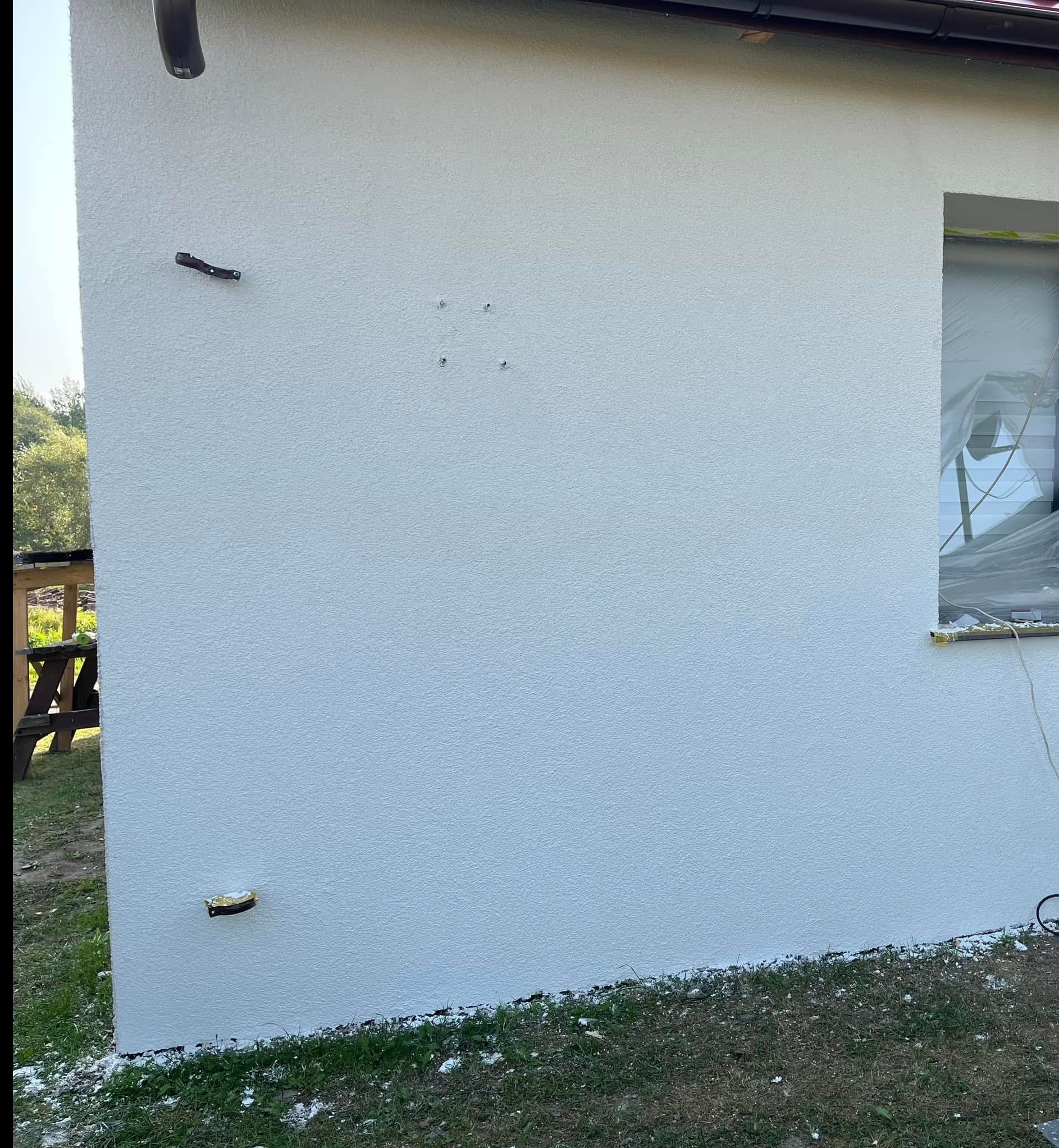 Biała, świeżo otynkowana ściana zewnętrzna domu z widocznymi elementami instalacji i zabezpieczonym oknem. Tekstura tynku barankowego.