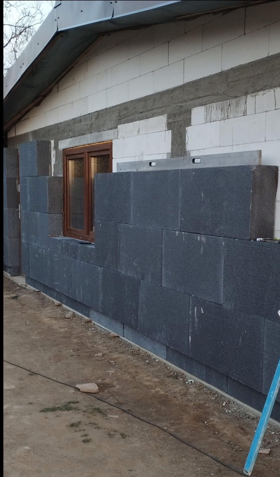 Ocieplanie budynku: ciemne bloki styropianu układane na ścianie z białego bloczka, okno w drewnianej ramie, poziomica na górze. Prace w toku.