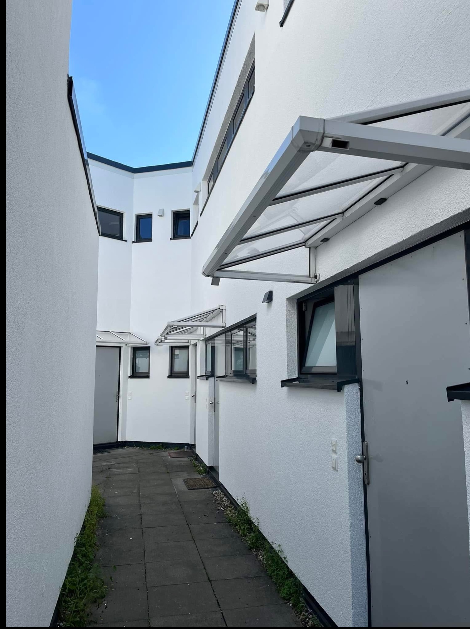 Białe budynki w wąskim przejściu, z szarymi drzwiami i daszkami, widok z dołu w kierunku nieba. Minimalistyczna architektura z wyraźnymi liniami i kątami.