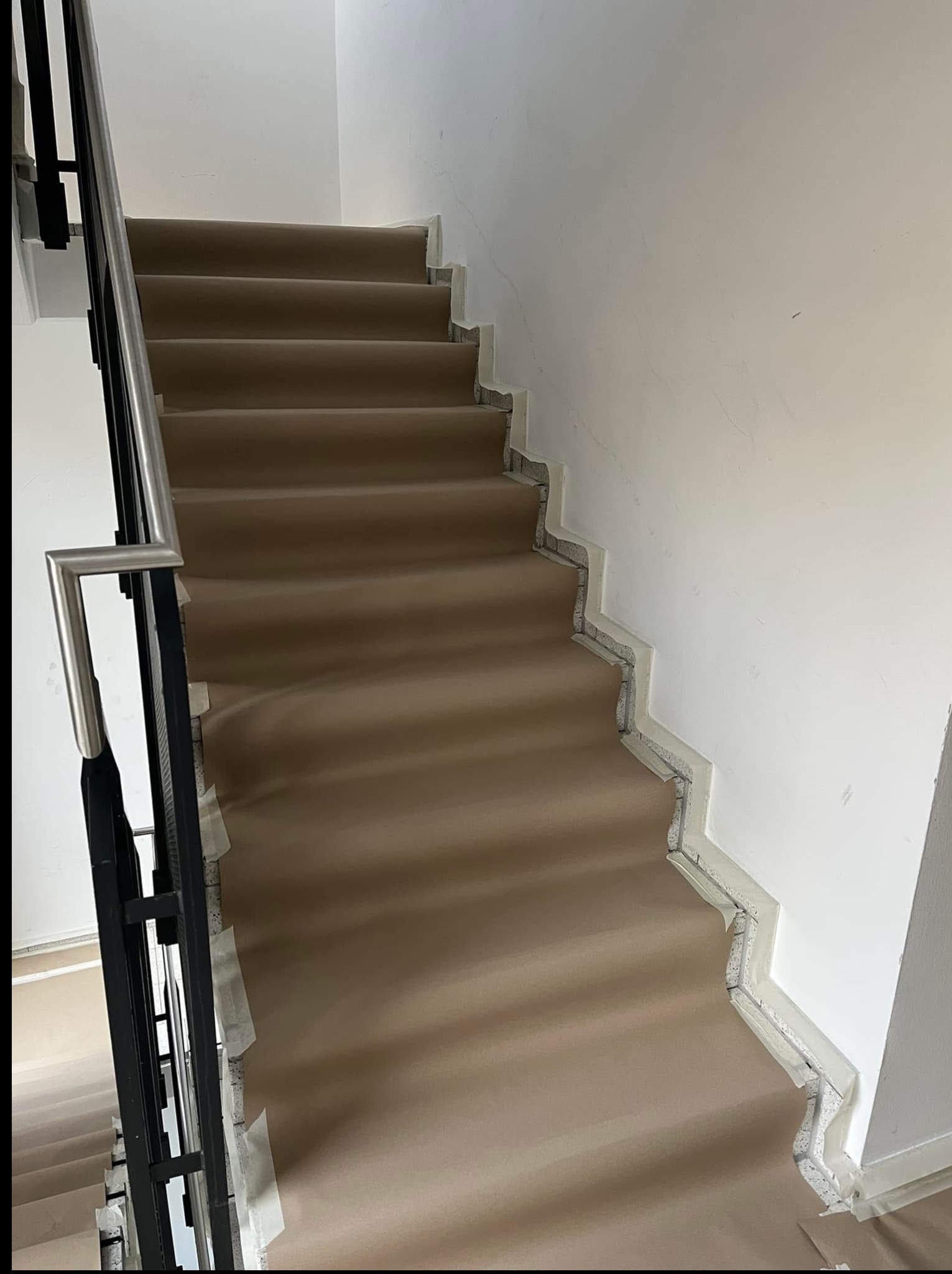 Wykończone schody z beżową wykładziną, zabezpieczone taśmą malarską przy ścianie i poręczy. Widok z góry na dół, skupienie na detalu wykończenia.