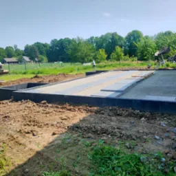 Przygotowany teren pod fundament z ułożonym obrzeżem, podsypką i płytami na piasku, widok na ogród i dom w tle, słoneczny dzień.