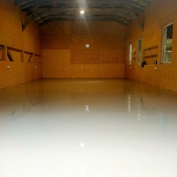 Kazare Quality Krzysztof Zaremba - Wnętrze budynku z płyt OSB z wylewką samopoziomującą na podłodze. Widoczne okna i drewniana konstrukcja dachu. Świeżo wylana, gładka powierzchnia podłogi.