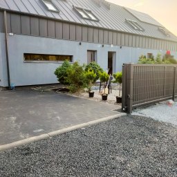 Kazare Quality Krzysztof Zaremba - Nowoczesny dom z szarą elewacją, metalowym ogrodzeniem i brukowanym podjazdem. Widoczne tuje w donicach i narzędzia ogrodnicze.