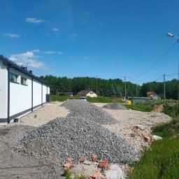 Kazare Quality Krzysztof Zaremba - Kupa gruzu i kamieni na placu budowy obok białego budynku. W tle zieleń drzew i fragmenty zabudowań. Niebieskie niebo z delikatnymi obłokami.