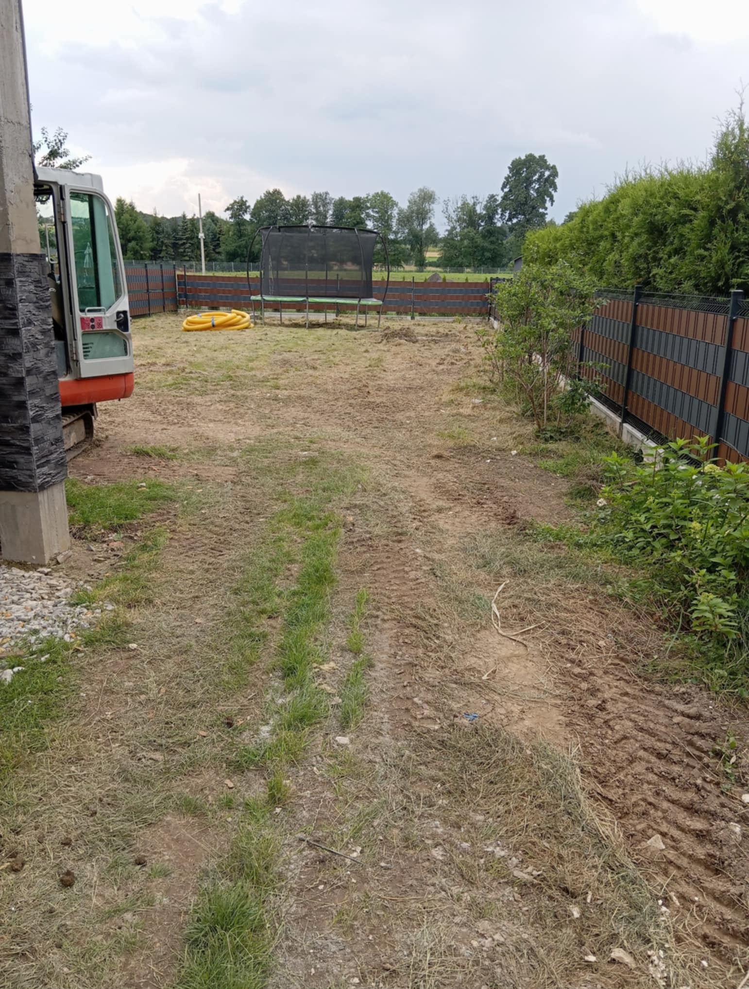 Teren po robotach ziemnych z widocznym ogrodzeniem panelowym, częściowo zarośnięty trawą, z widocznymi śladami opon. W tle trampolina.