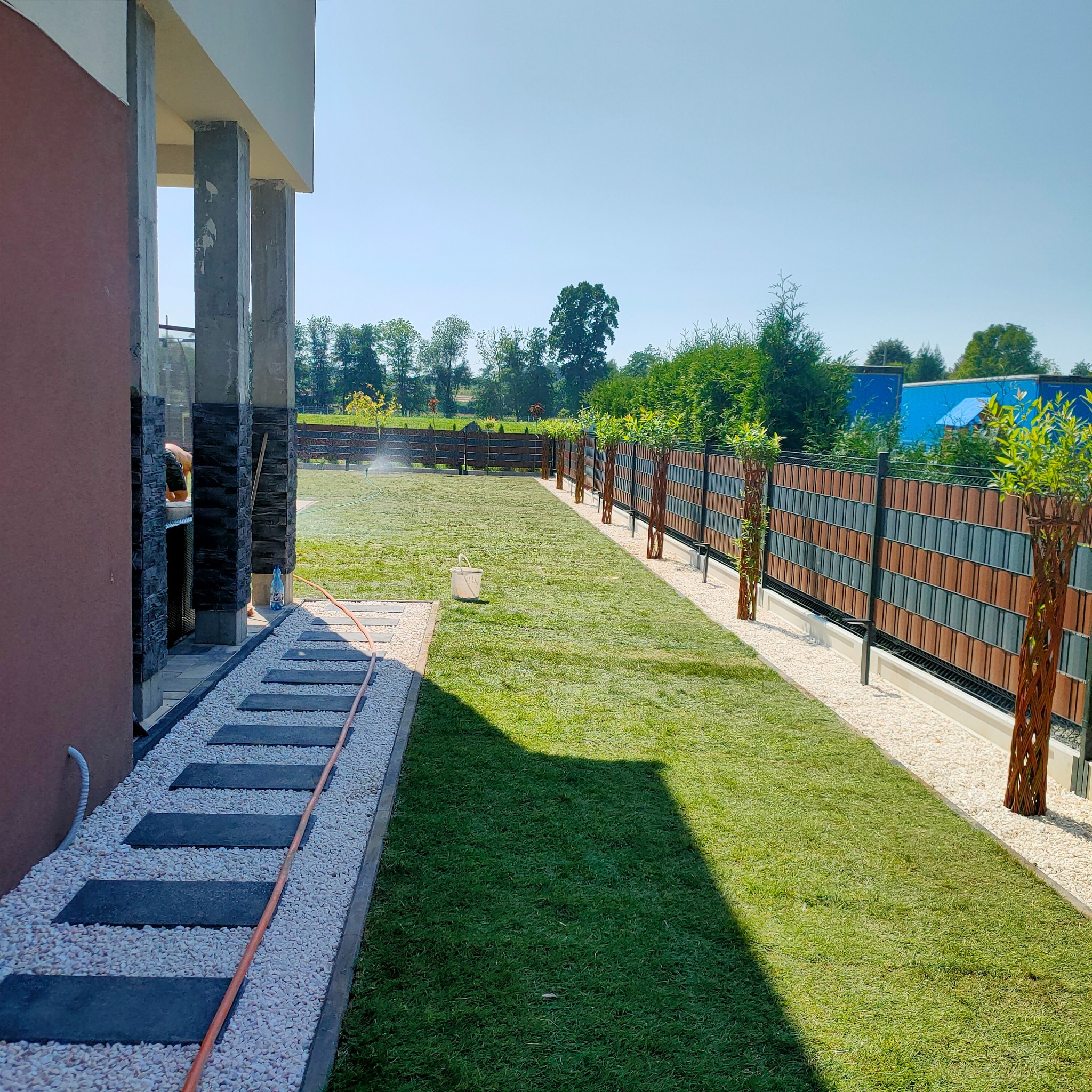 Nowoczesny ogród z panelowym ogrodzeniem w kolorze drewna i szarości, trawnikiem, ścieżką z kamieni i nawadnianiem. W tle zieleń drzew i błękitne niebo.