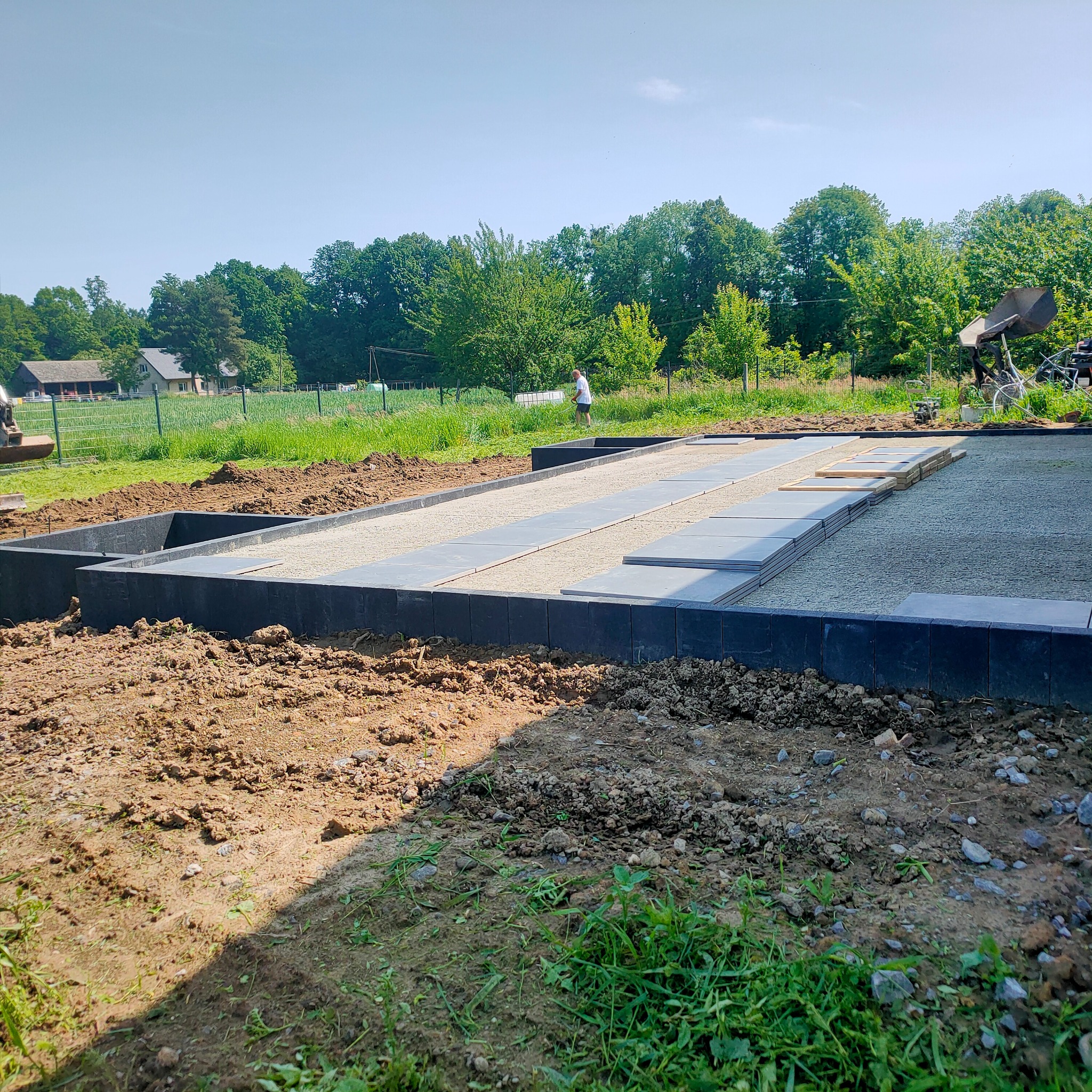 Przygotowany teren pod fundament z ułożonym obrzeżem, podsypką i płytami na piasku, widok na ogród i dom w tle, słoneczny dzień.