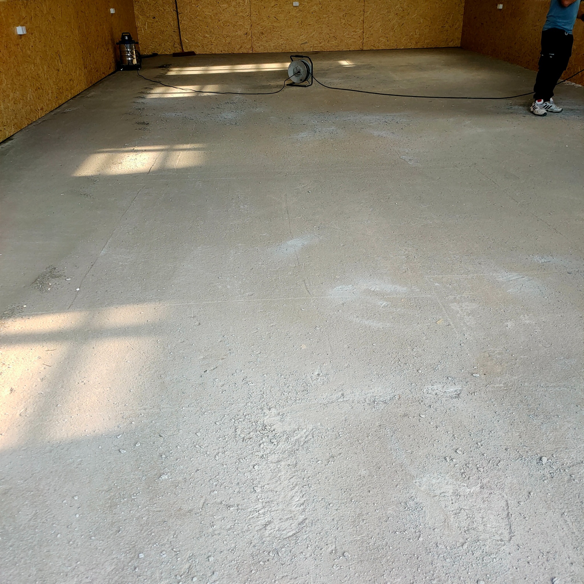 Wnętrze w trakcie wykończenia: surowy beton na podłodze, ściany z płyt OSB, widoczny odkurzacz przemysłowy i zwinięty kabel.