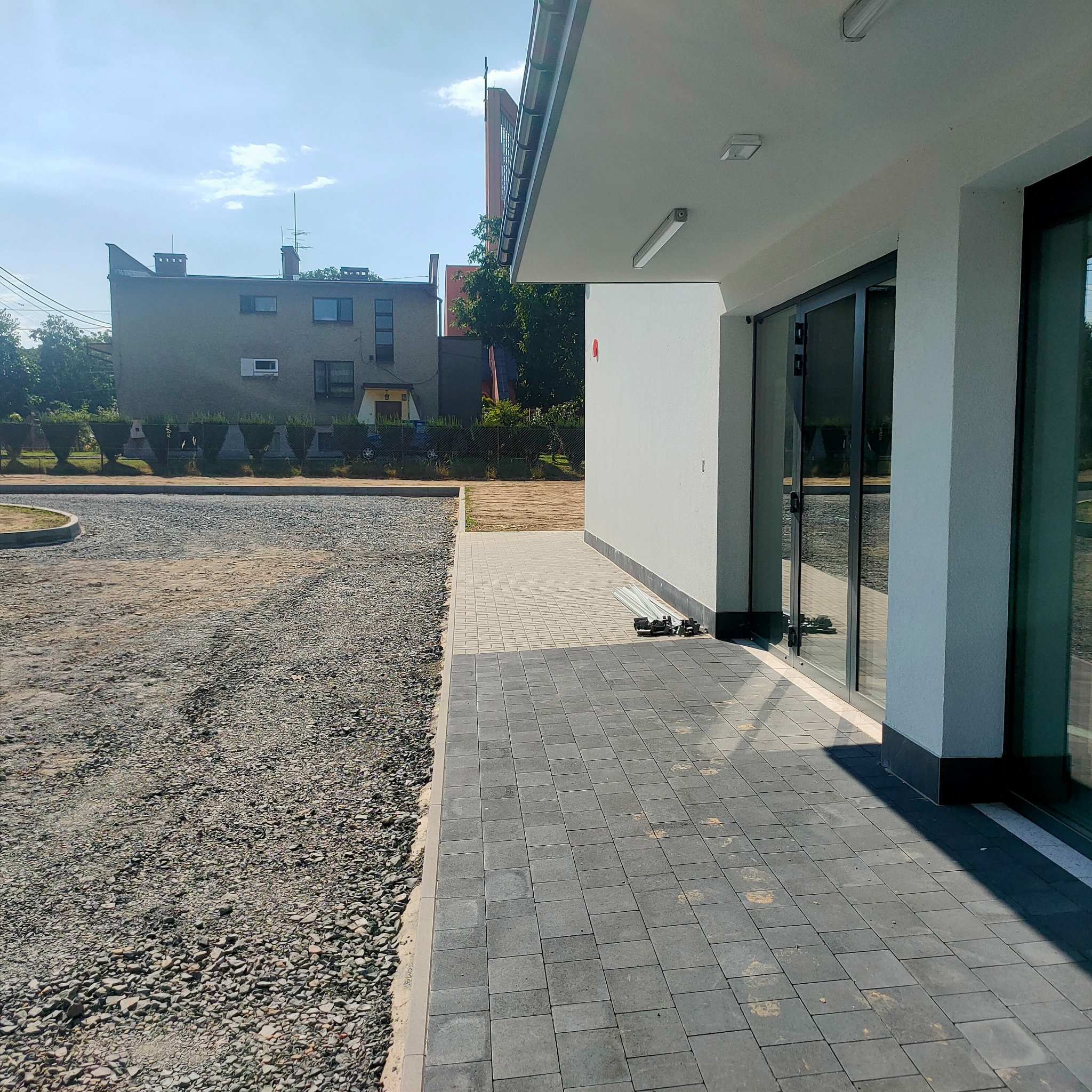 Ułożona kostka brukowa przy budynku z widocznym podłożem z kruszywa. Elementy konstrukcyjne ogrodzenia leżące obok budynku.