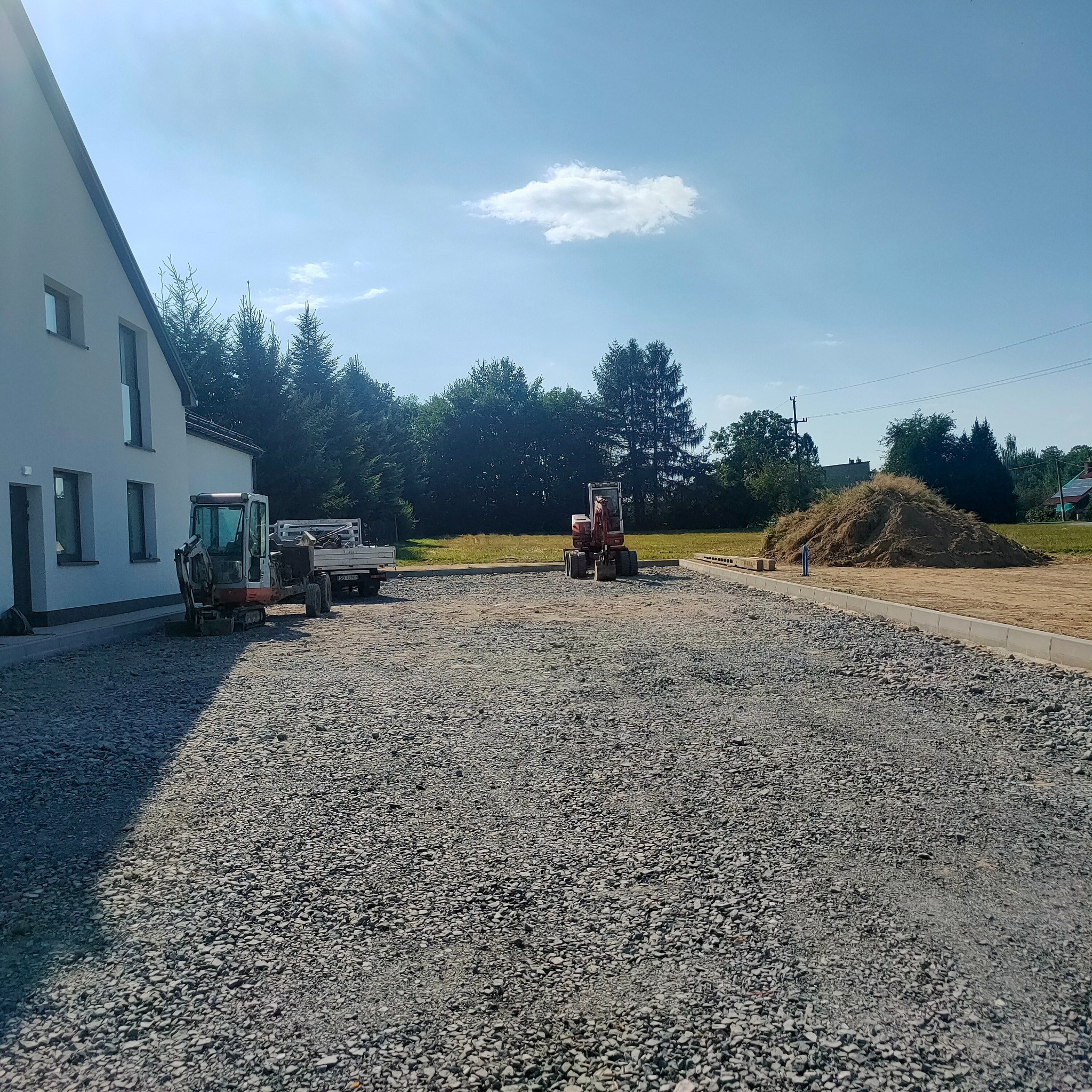 Teren po utwardzeniu gruzem, z widocznymi dwoma małymi koparkami i stertą ziemi w tle, obok budynku. Słoneczny dzień.
