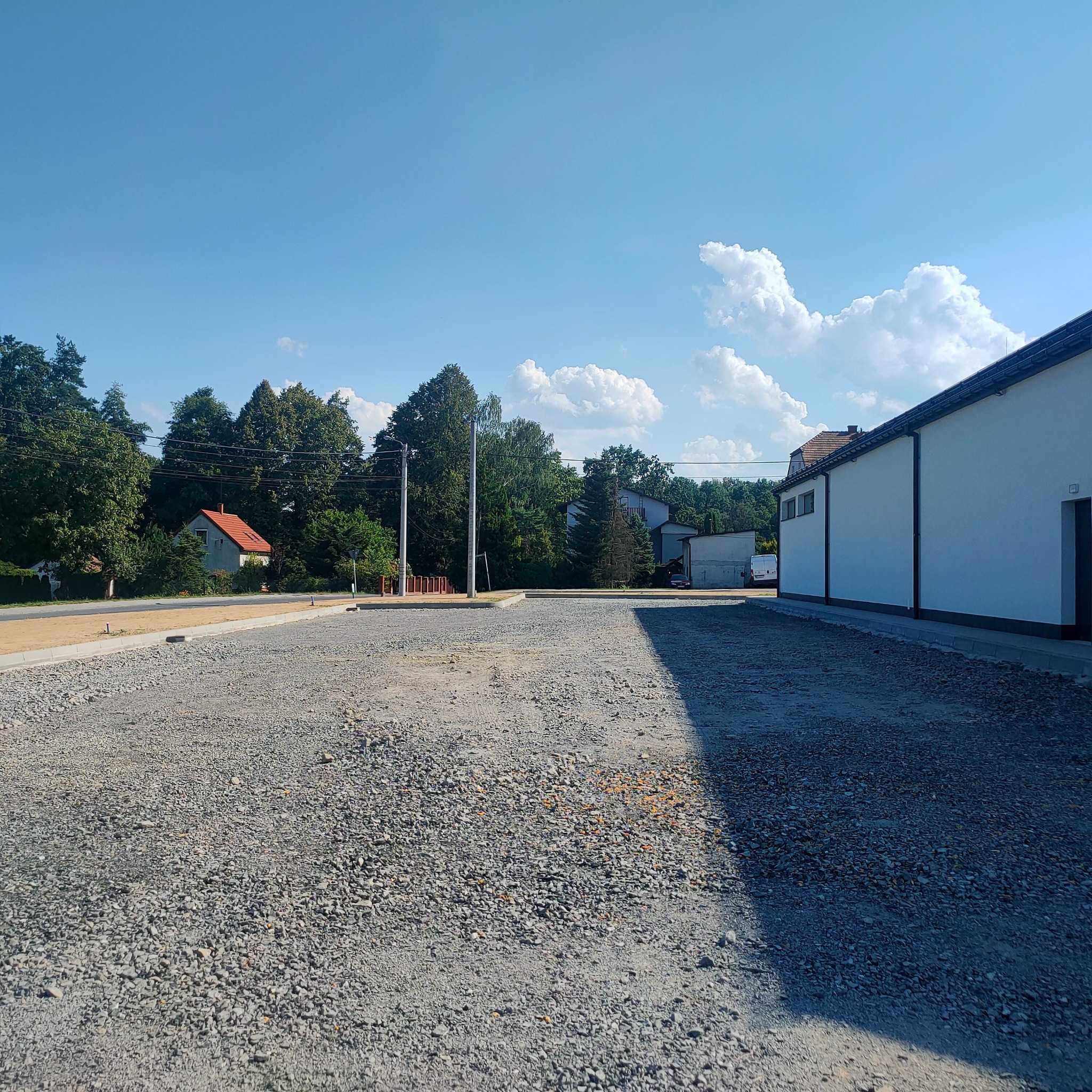 Utwardzony teren z szarą nawierzchnią z kamienia, obok budynek z białymi ścianami, w tle zieleń drzew i błękitne niebo z chmurami.