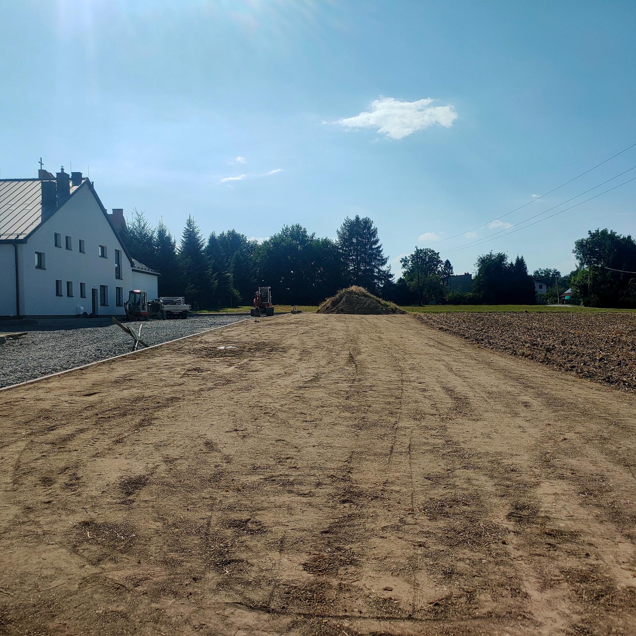 Przygotowany teren pod budowę drogi dojazdowej, widoczny gruz, ubita ziemia i koparka w tle. W oddali biały budynek z ciemnym dachem.