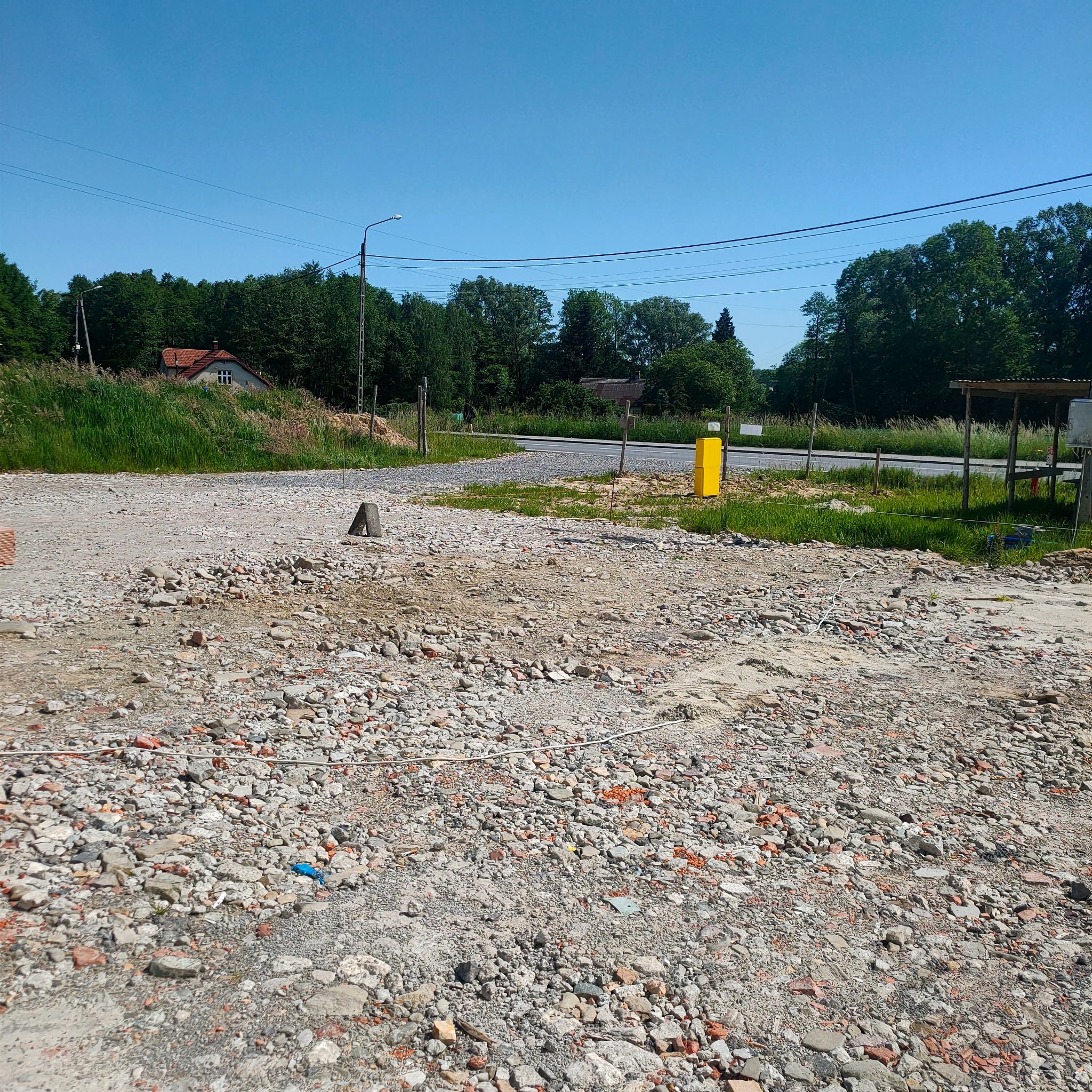 Teren po utwardzeniu gruzem i kamieniami, widoczny fragment drogi, drzewa w tle. Przygotowanie terenu pod budowę lub utwardzenie placu.