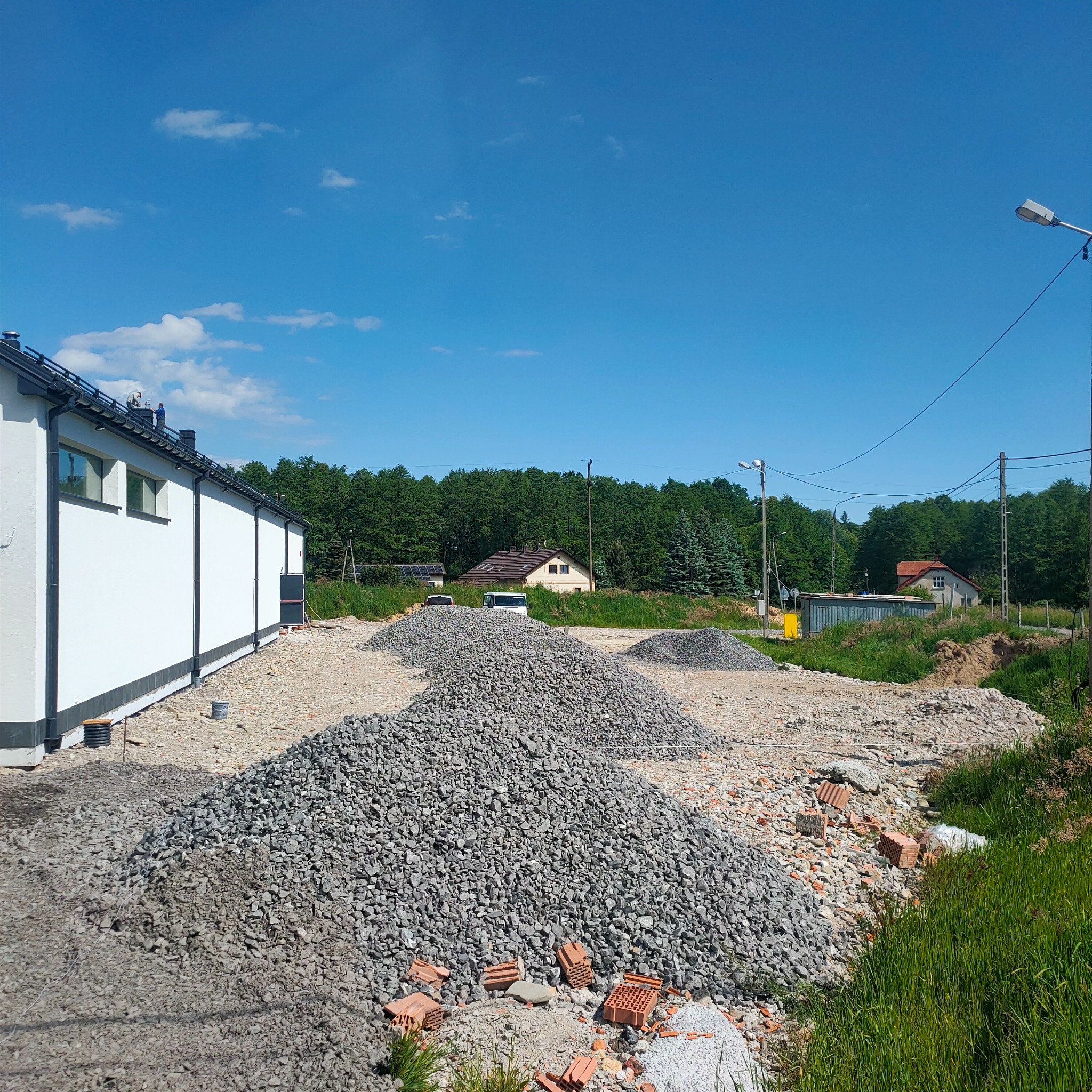 Kupa gruzu i kamieni na placu budowy obok białego budynku. W tle zieleń drzew i fragmenty zabudowań. Niebieskie niebo z delikatnymi obłokami.