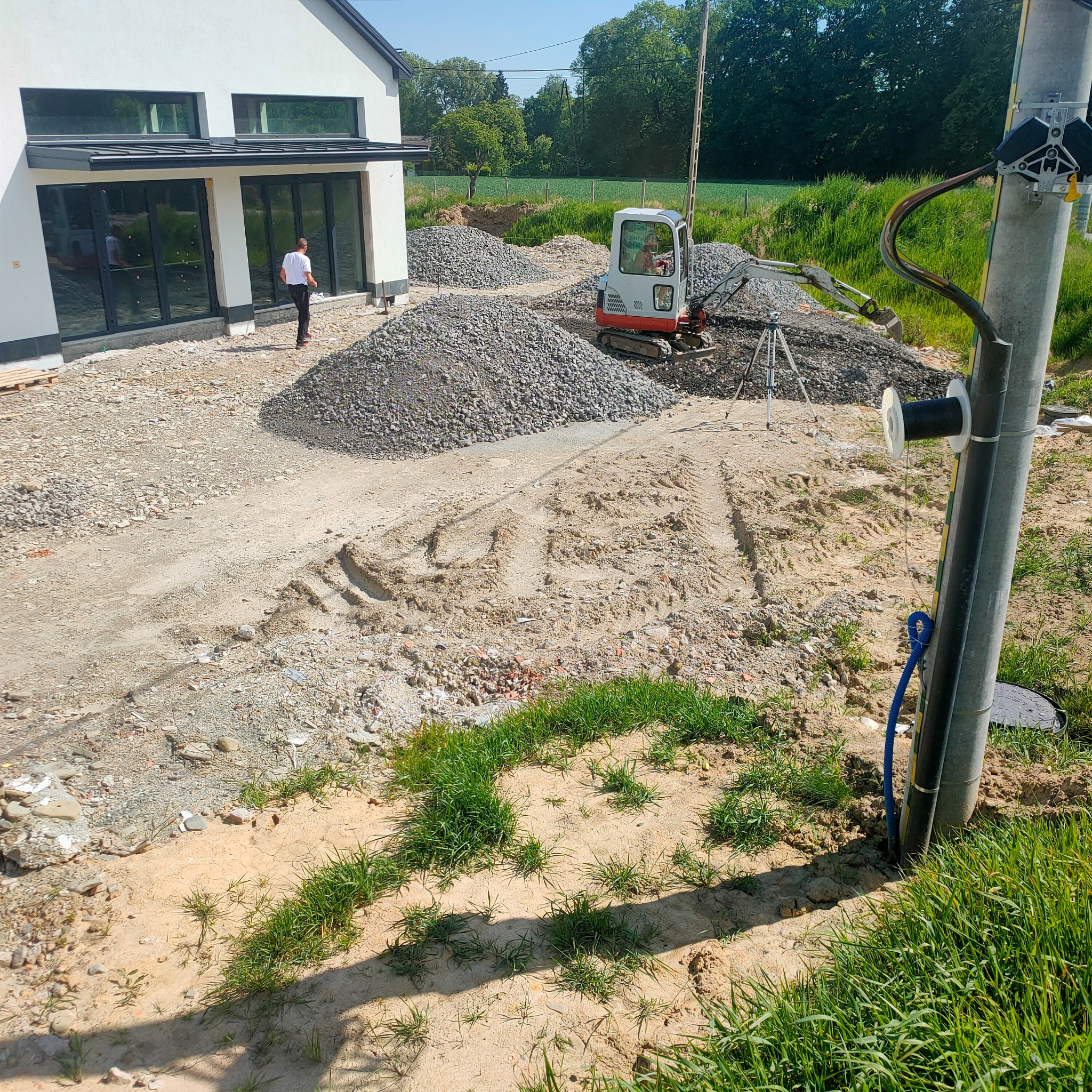 Mini koparka przygotowuje teren pod budowę domu jednorodzinnego. Widoczne sterty gruzu, wykopana ziemia i fundamenty budynku w tle, słoneczny dzień.