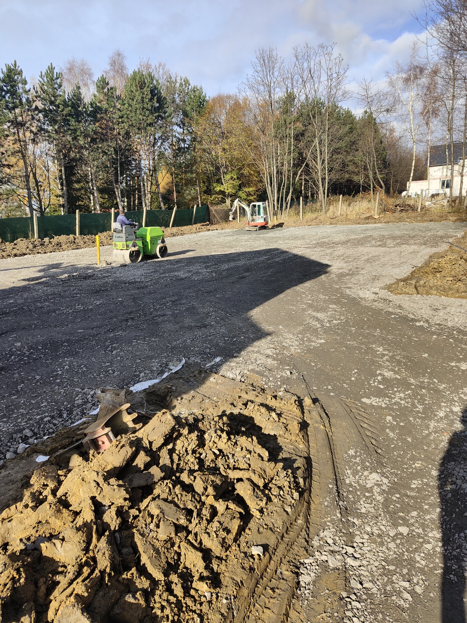 Utwardzanie terenu pod budowę: walec drogowy w akcji, ubijający gruz i ziemię. W tle widoczna mała koparka i drzewa.