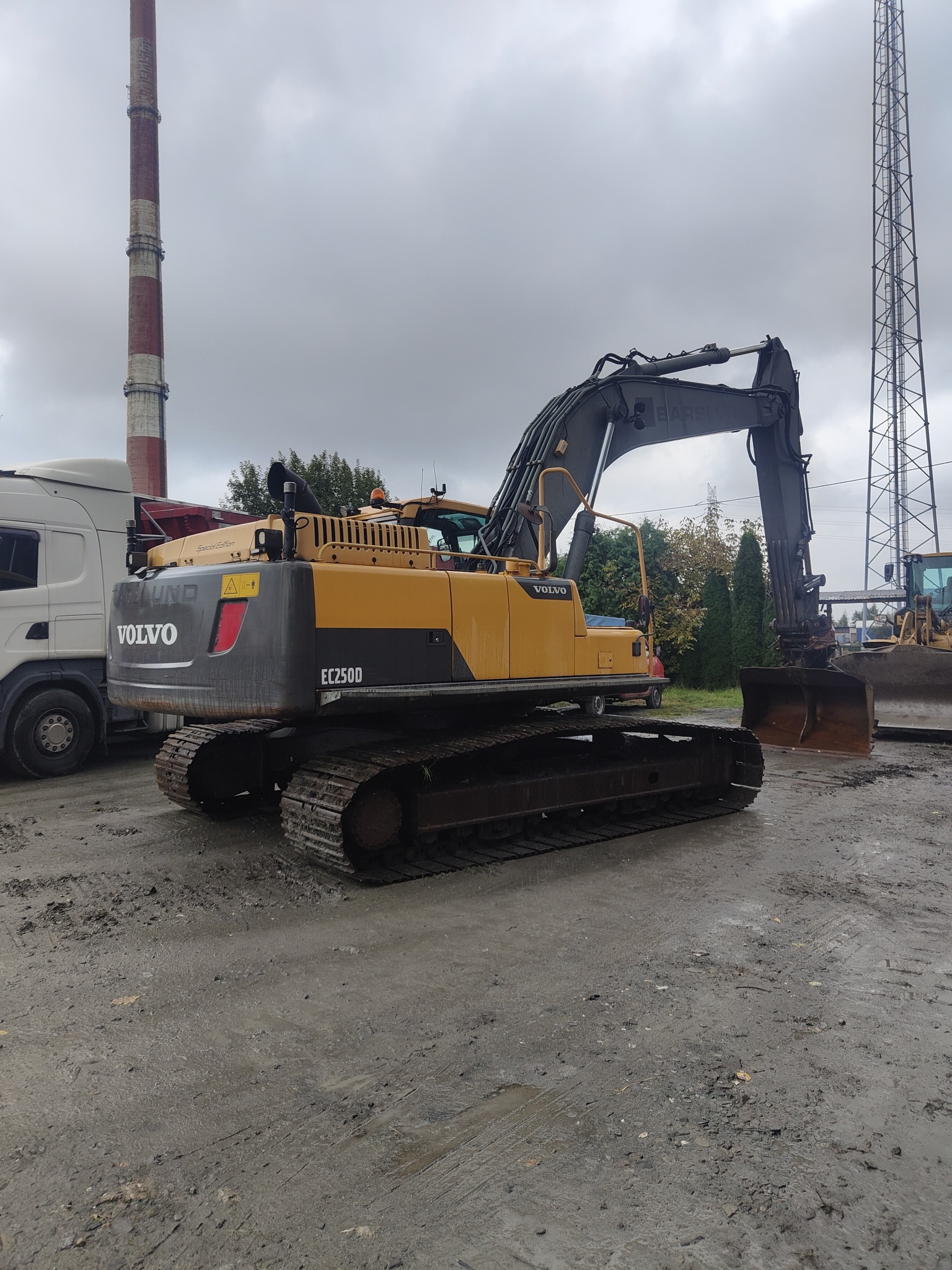 Żółta koparka Volvo EC250D na placu budowy, z widocznym kominem i wieżą transmisyjną w tle, sugerująca prace ziemne lub rozbiórkowe.