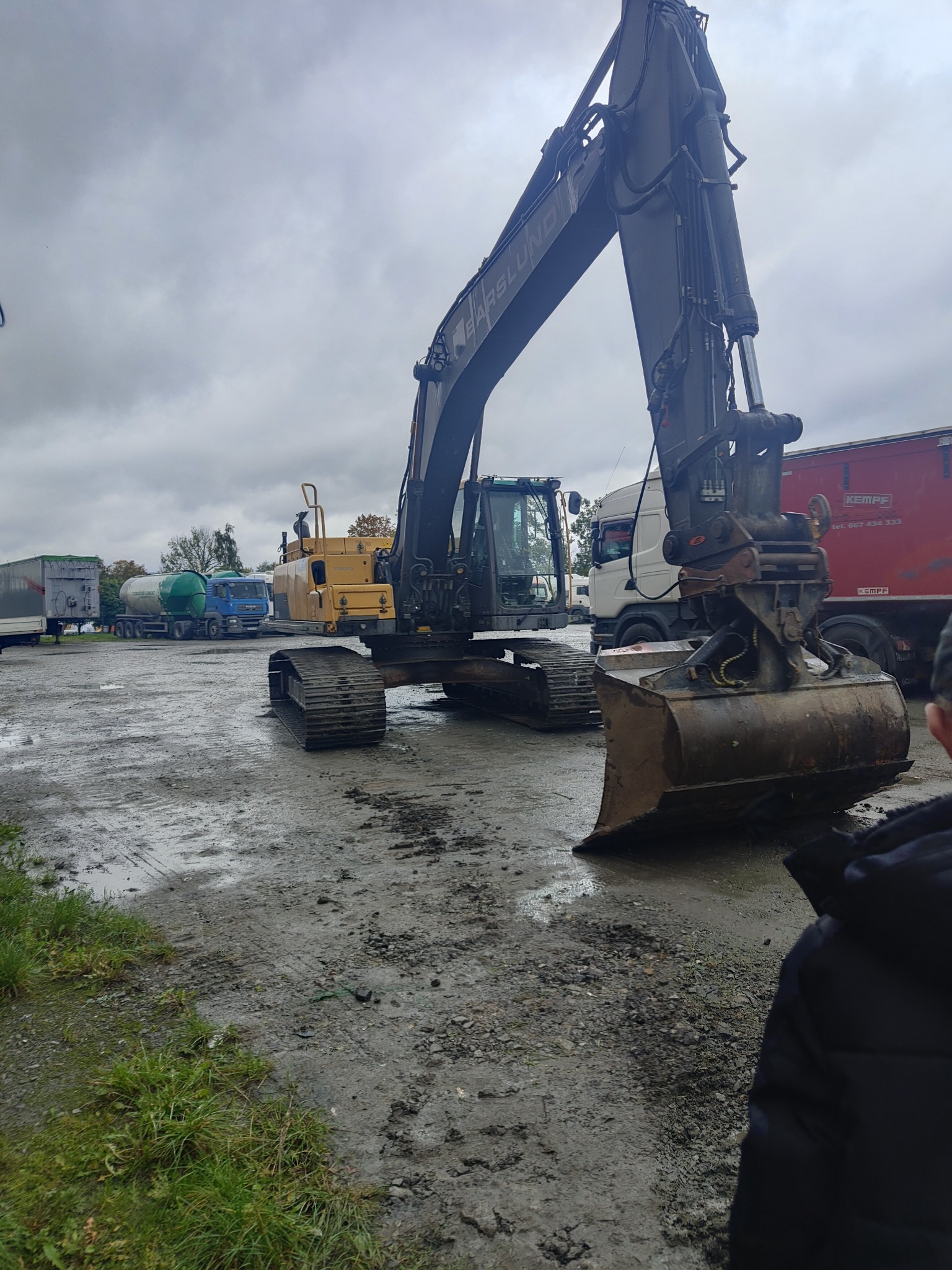 Żółto-czarna koparka gąsienicowa marki Barslund na błotnistym terenie, z widocznymi ciężarówkami w tle pod pochmurnym niebem. Sprzęt budowlany w trakcie pracy.