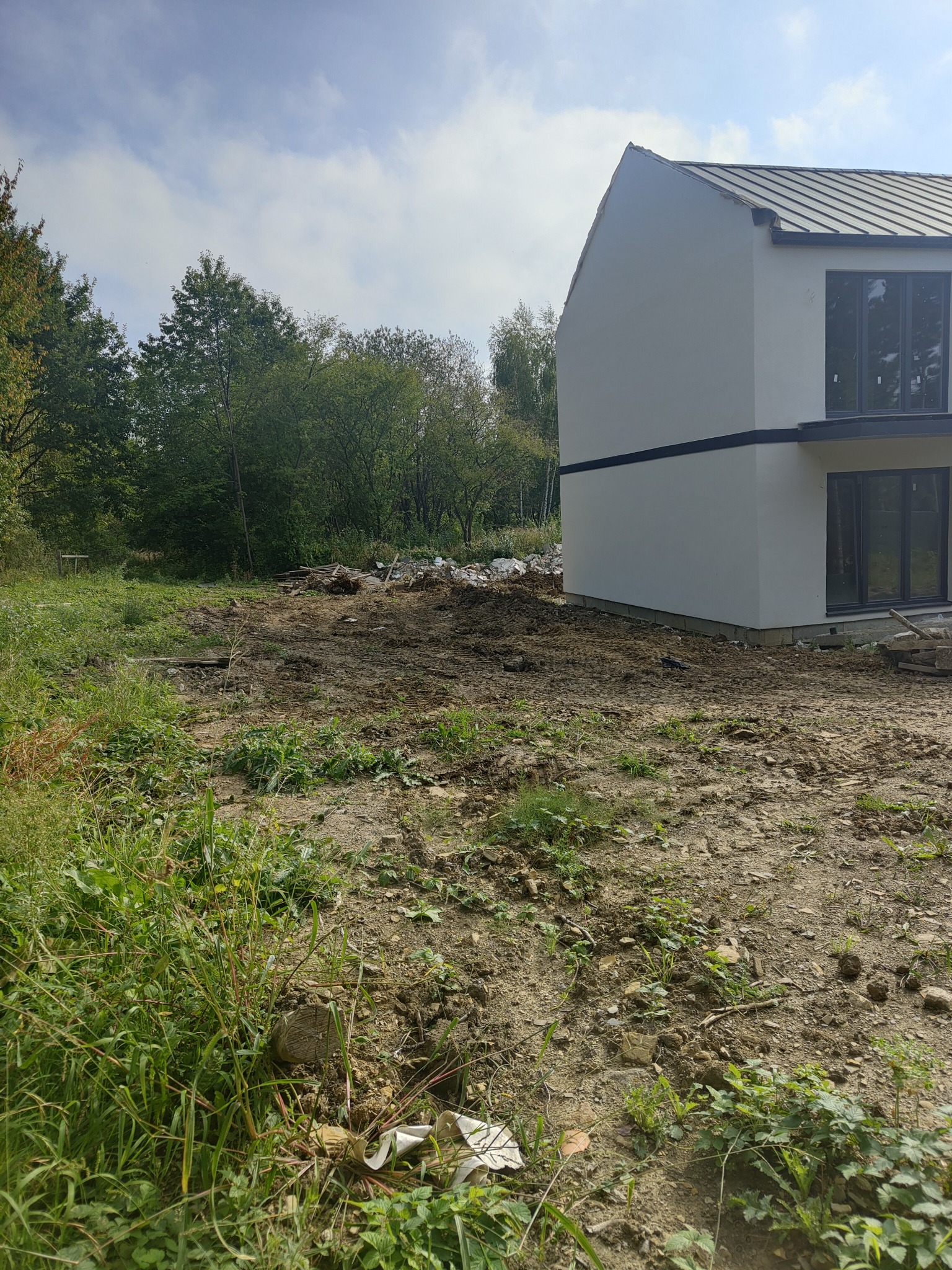 Dwu kondygnacyjny dom z szarą blachą na dachu, widoczny z boku na tle zieleni. Teren wokół domu częściowo wyrównany, z pozostałościami gruzu i ziemi.