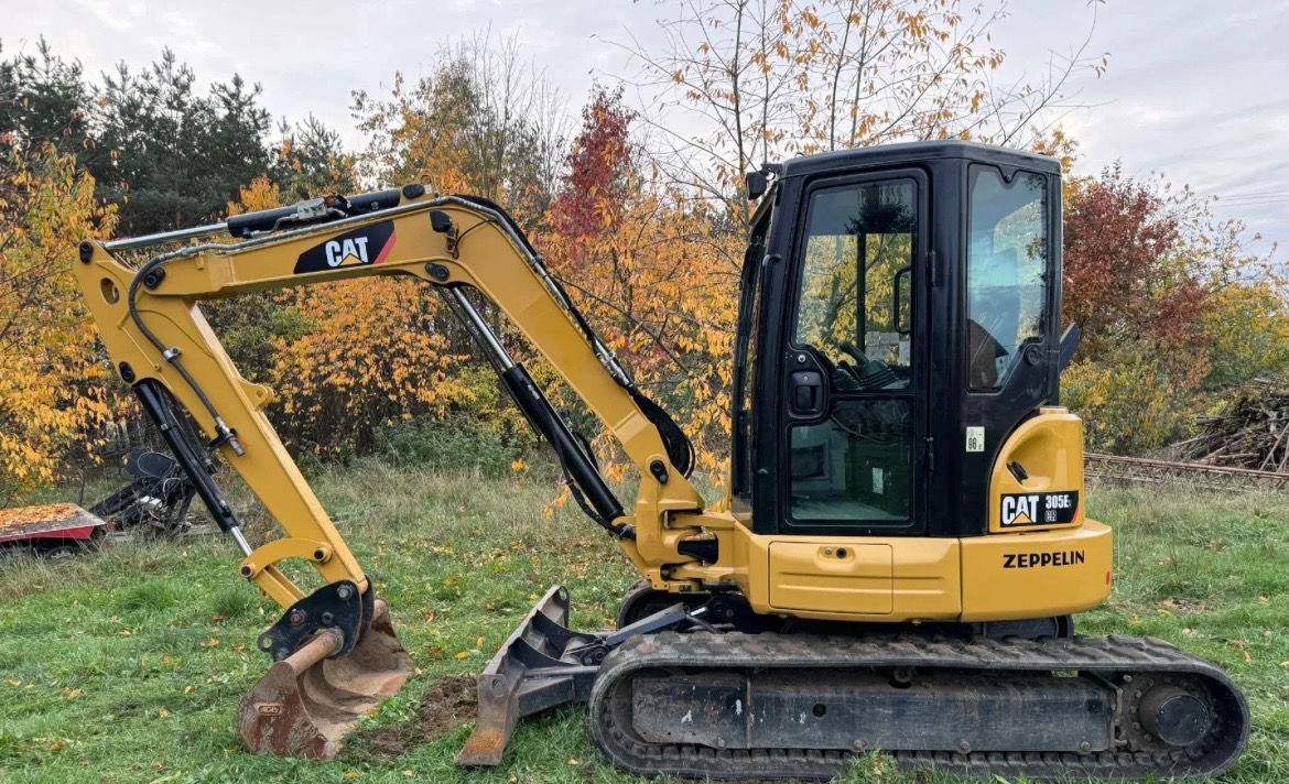 Żółta koparka CAT 305E na trawie, z łyżką w ziemi. Widoczne jesienne drzewa w tle. Sprzęt budowlany w trakcie prac ziemnych.