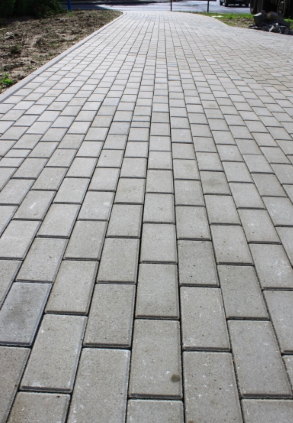 Długa, wybrukowana ścieżka z szarych, prostokątnych kostek, biegnąca w perspektywie z bliska do oddali, z zielenią i fragmentem pojazdu w tle.
