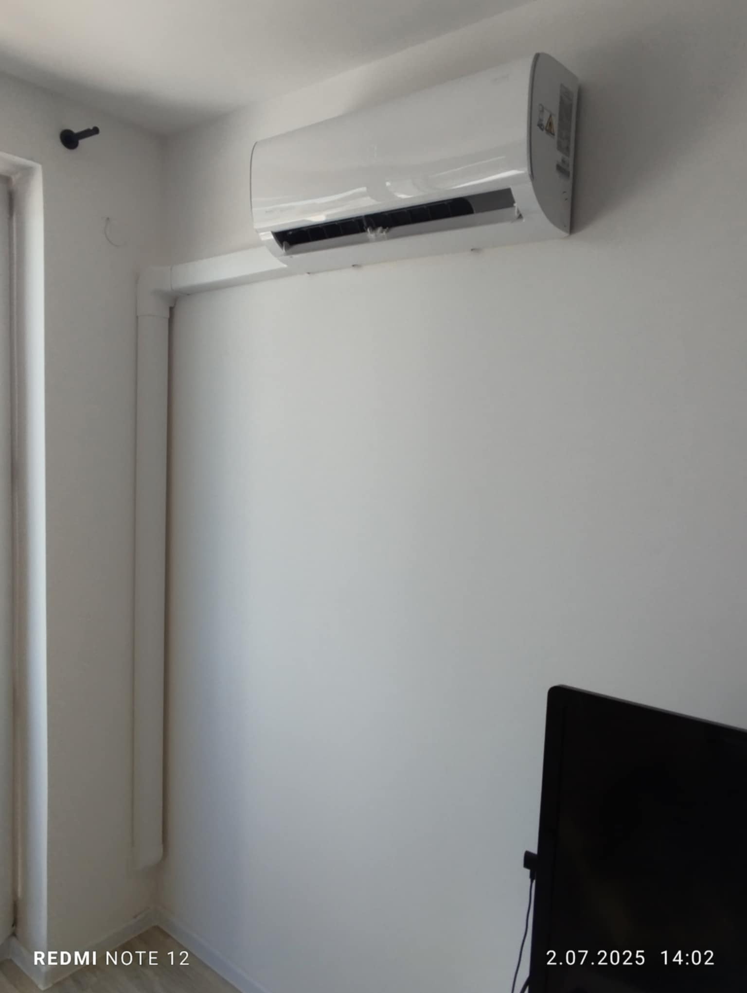 Biały klimatyzator na ścianie w pomieszczeniu, widoczne rury instalacyjne, fragment telewizora w rogu. Minimalistyczny design, neutralne tło.