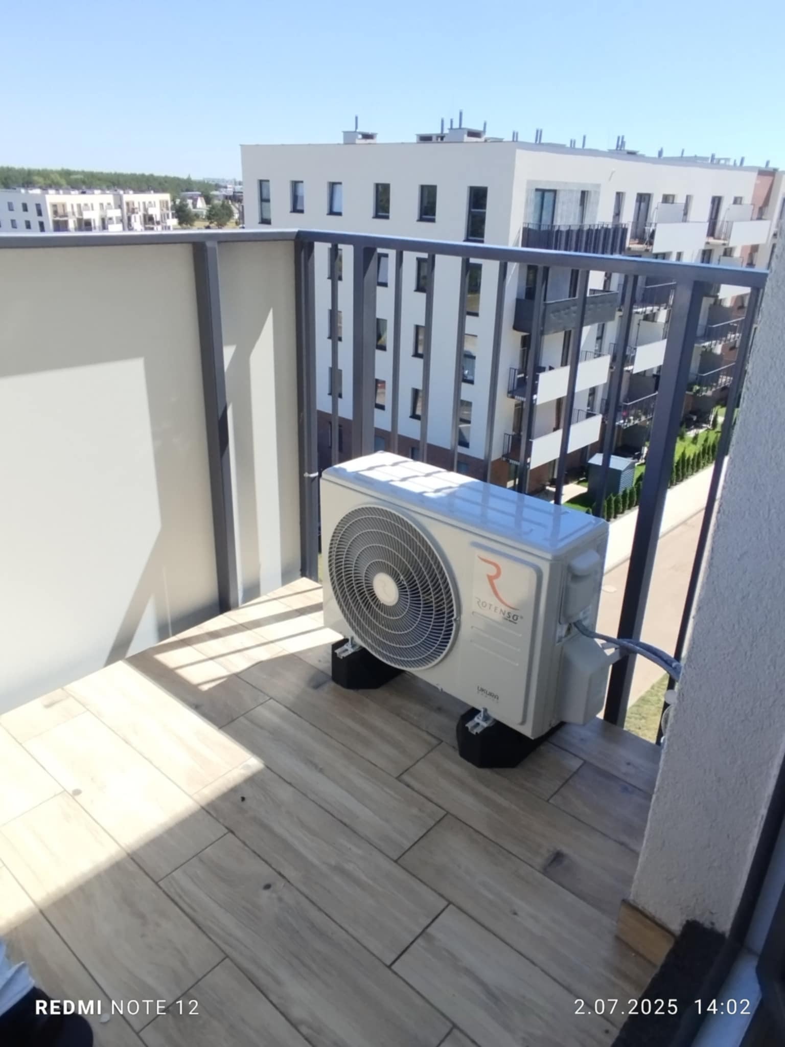 Zamontowana jednostka zewnętrzna klimatyzacji Rotenso na balkonie w bloku mieszkalnym. Widoczne elementy montażowe i przewody. Jasne otoczenie, wysoki budynek w tle.