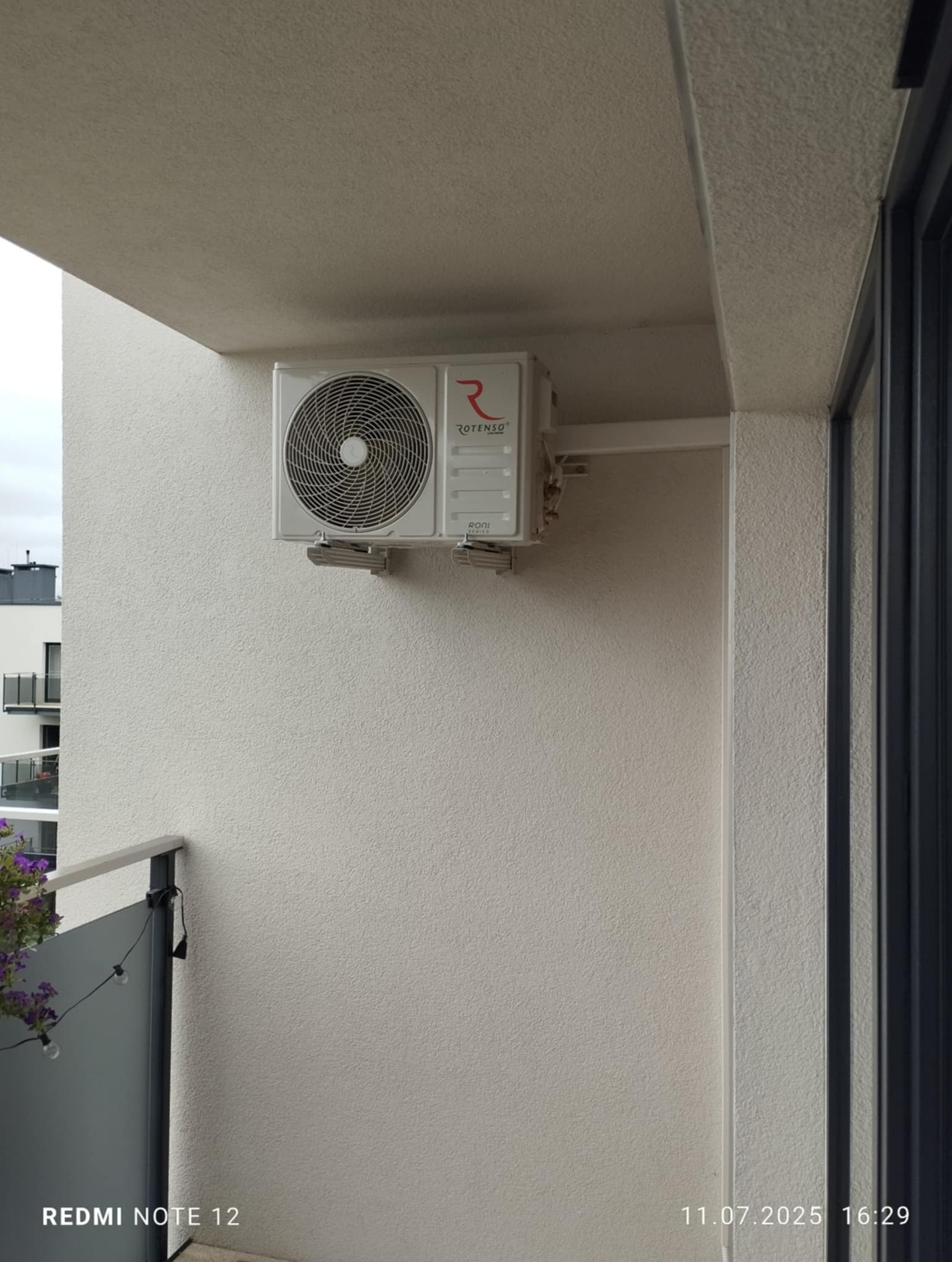 Zamontowany klimatyzator Rotenso na balkonie nowoczesnego budynku. Widoczne mocowania i fragment balustrady. Ściana o jasnej, chropowatej fakturze.