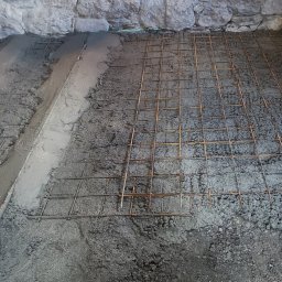 KazDachy F. H. U - Przygotowanie pod wylewkę: zbrojenie z siatki stalowej na warstwie betonu, widoczne ściany z kamienia w surowym stanie, zielony laser poziomujący.