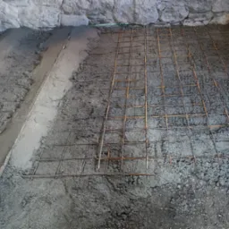 Przygotowanie pod wylewkę: zbrojenie z siatki stalowej na warstwie betonu, widoczne ściany z kamienia w surowym stanie, zielony laser poziomujący.