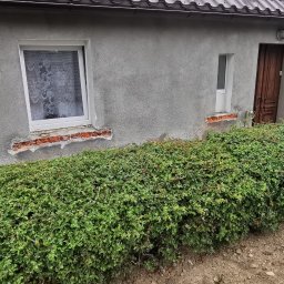 KazDachy F. H. U - Szary budynek w trakcie docieplania. Widoczne fragmenty styropianu, pistolet do pianki montażowej, okna z białymi ramami. Częściowo ocieplona elewacja.