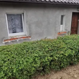 Szary budynek w trakcie docieplania. Widoczne fragmenty styropianu, pistolet do pianki montażowej, okna z białymi ramami. Częściowo ocieplona elewacja.