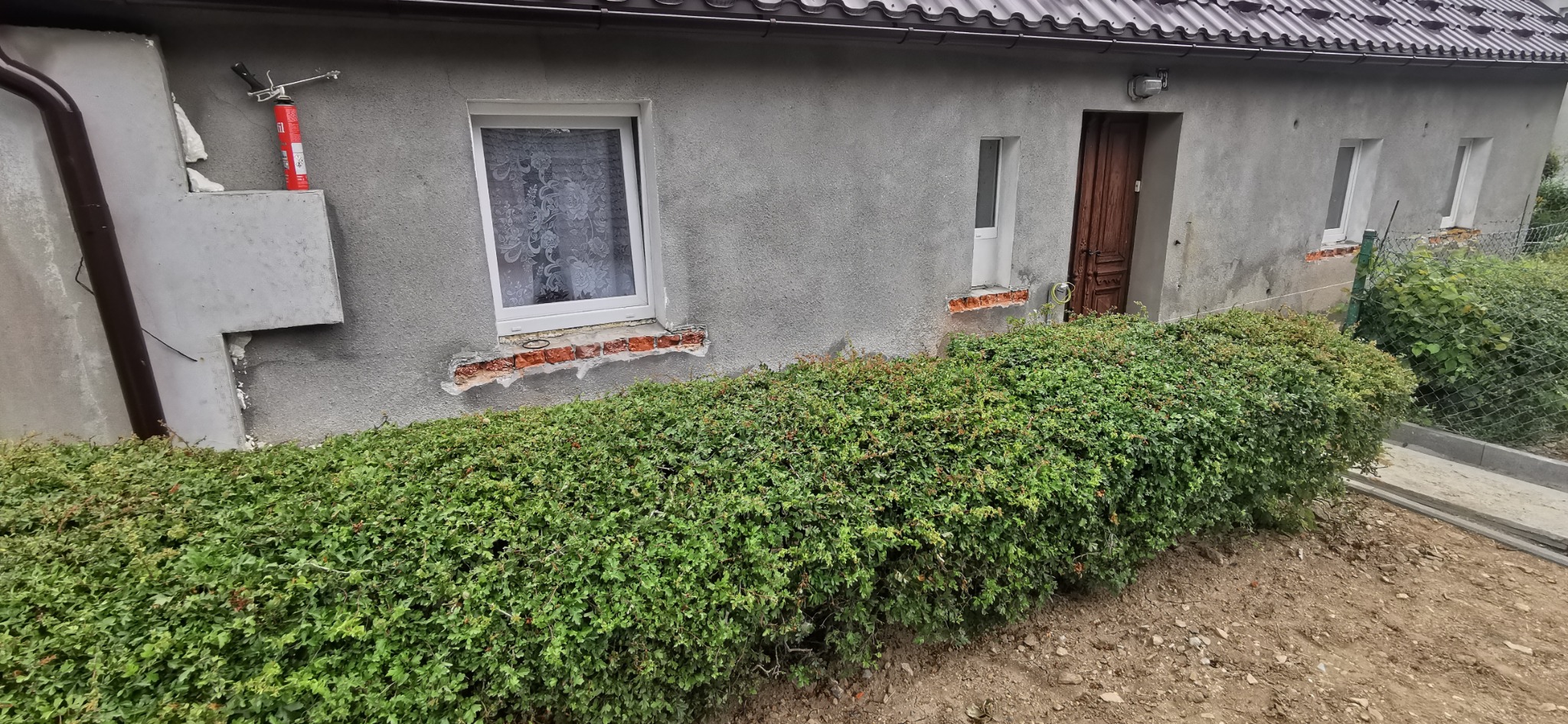 Szary budynek w trakcie docieplania. Widoczne fragmenty styropianu, pistolet do pianki montażowej, okna z białymi ramami. Częściowo ocieplona elewacja.