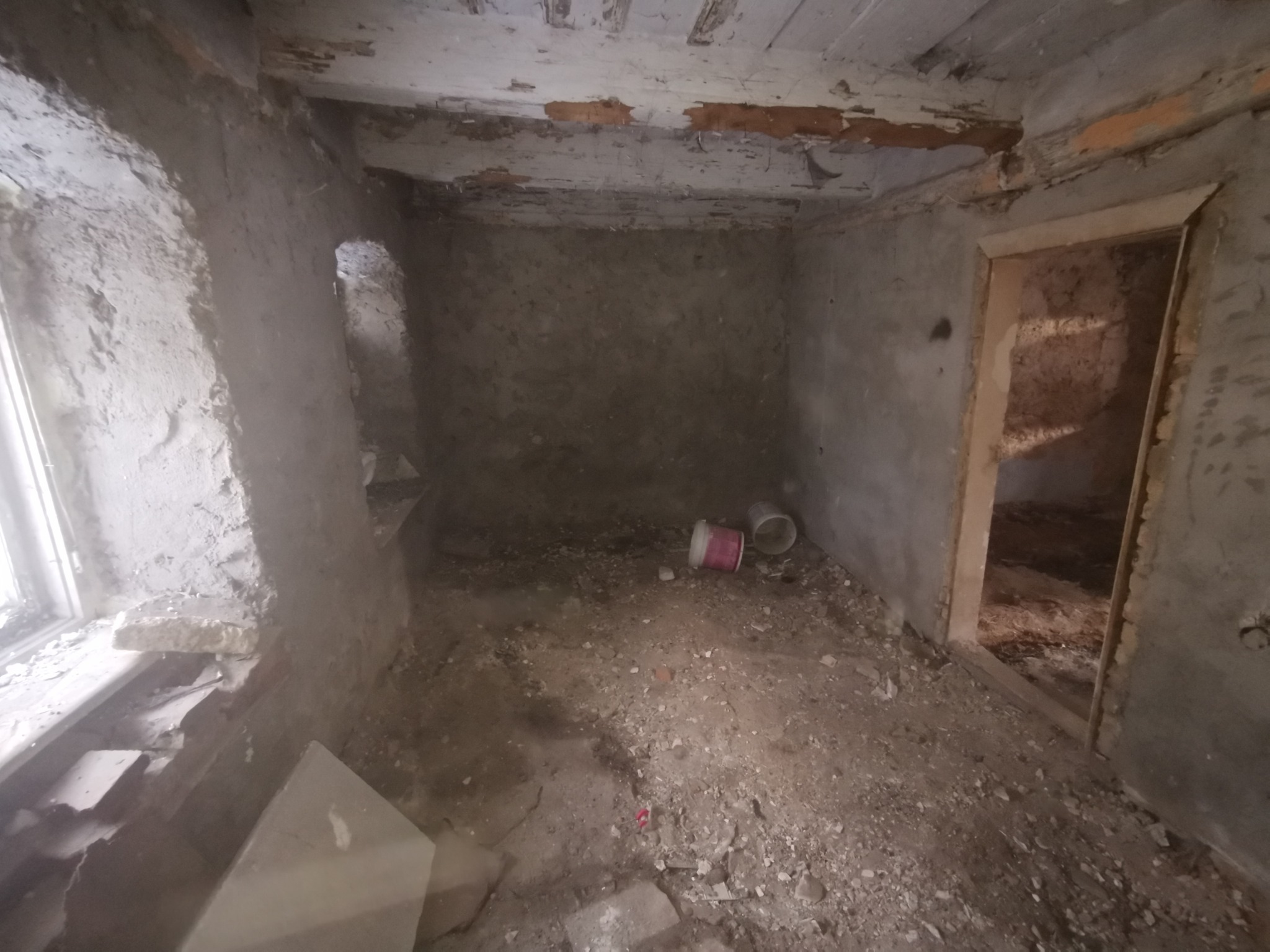 Wnętrze zaniedbanego pokoju przed remontem: surowe ściany, okno, gruz na podłodze, widoczne puszki po farbie. Elementy starej stolarki okiennej i drzwiowej.