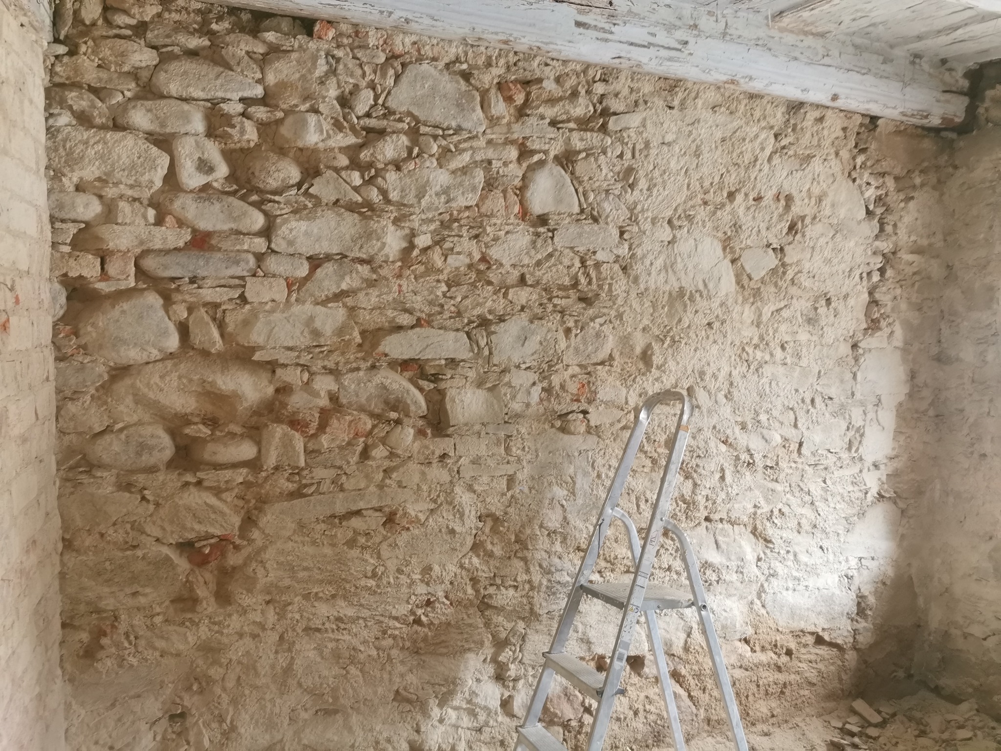 Przygotowanie ściany z kamienia pod tynkowanie. Widoczna drabina, oczyszczone kamienie i uzupełnione fugi. Surowy stan przed dalszymi pracami wykończeniowymi.