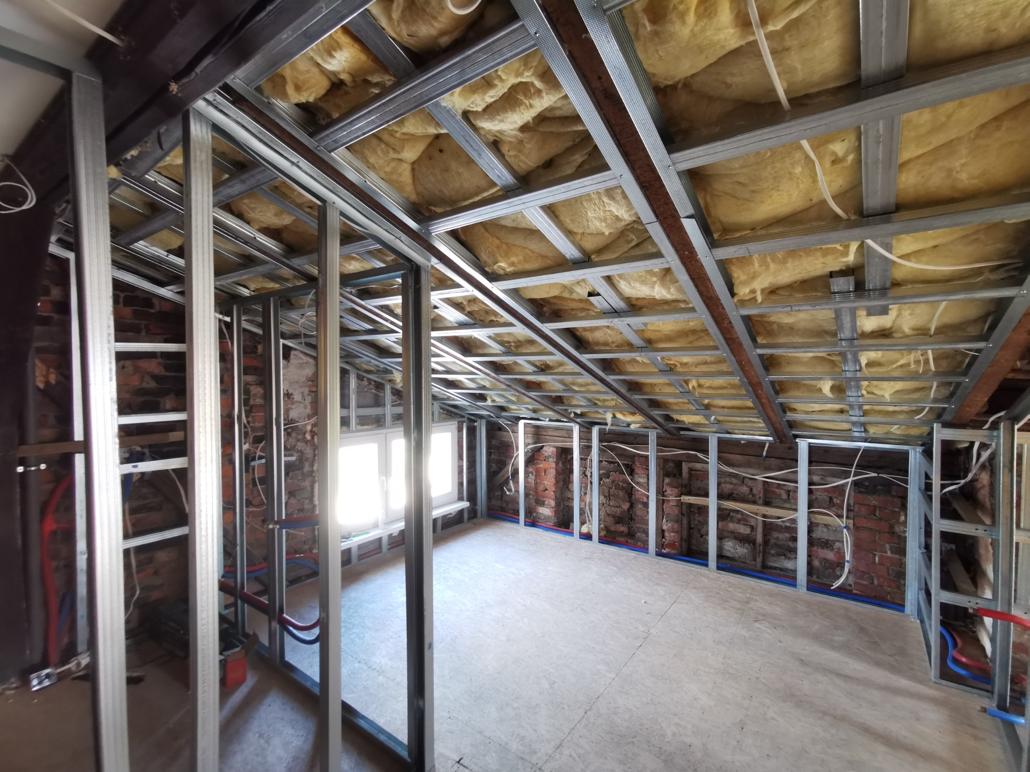 Adaptacja poddasza: widok na stelaż z płyt karton-gips, izolację między krokwiami i instalacje. Elementy z bliska, surowy stan, w tle okno.