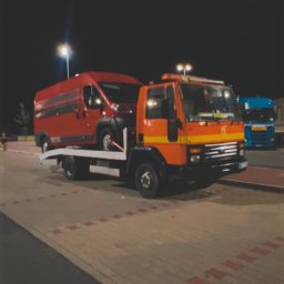 JOKER - Pomarańczowa laweta z czerwonym busem na platformie, nocą, na parkingu z kostki brukowej. W tle widoczny niebieski samochód ciężarowy.